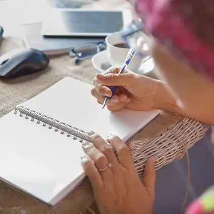 Persoon schrijft in een notitieboek aan tafel, omringd door computer en koffie, terwijl uitvaartwensen worden vastgelegd.